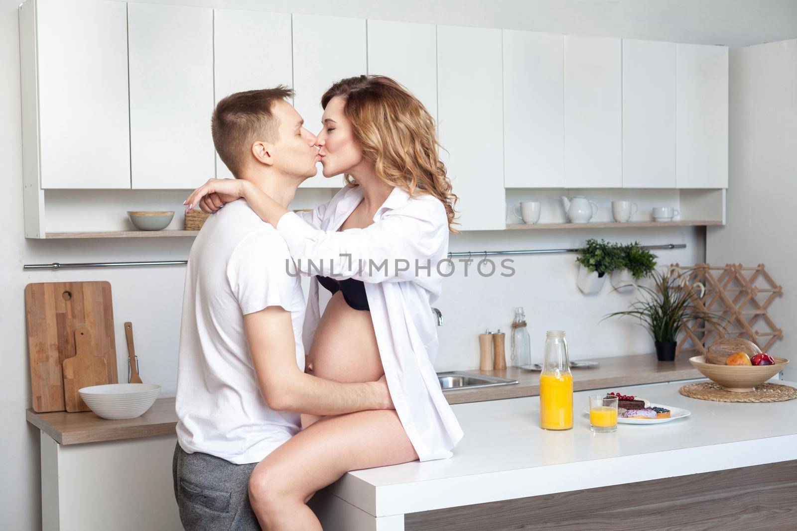 Lovestory. Young married couple embraces siting on table in kitchen. Husband hugs his pregnant wife, putting his hands on her big belly, parents enjoy each other and kissing with closed eyes.