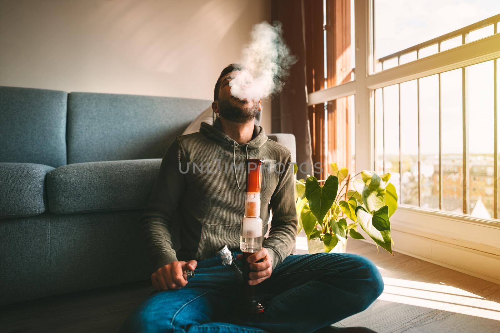 Man smoking bong and exhaling the smoke at home. Man smoking pot, medical marijuana or cannabis from a bong or water pipe. Cannabis and weed legalisation concept. High quality photo