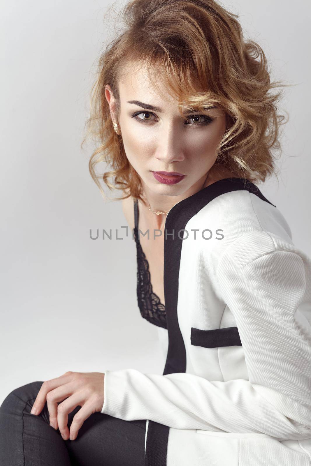 Portrait of hot young sexy model, posing in bra and undressing her white jacket and looking at camera. Studio shot, gray wall