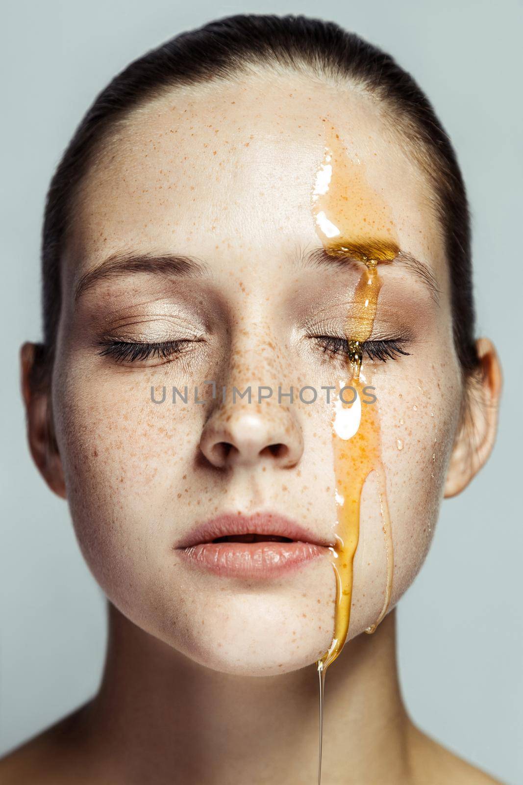 Portrait of beautiful young brunette woman with freckles and honey on face with closed eyes and serious face. indoor studio shot isolated on gray background.