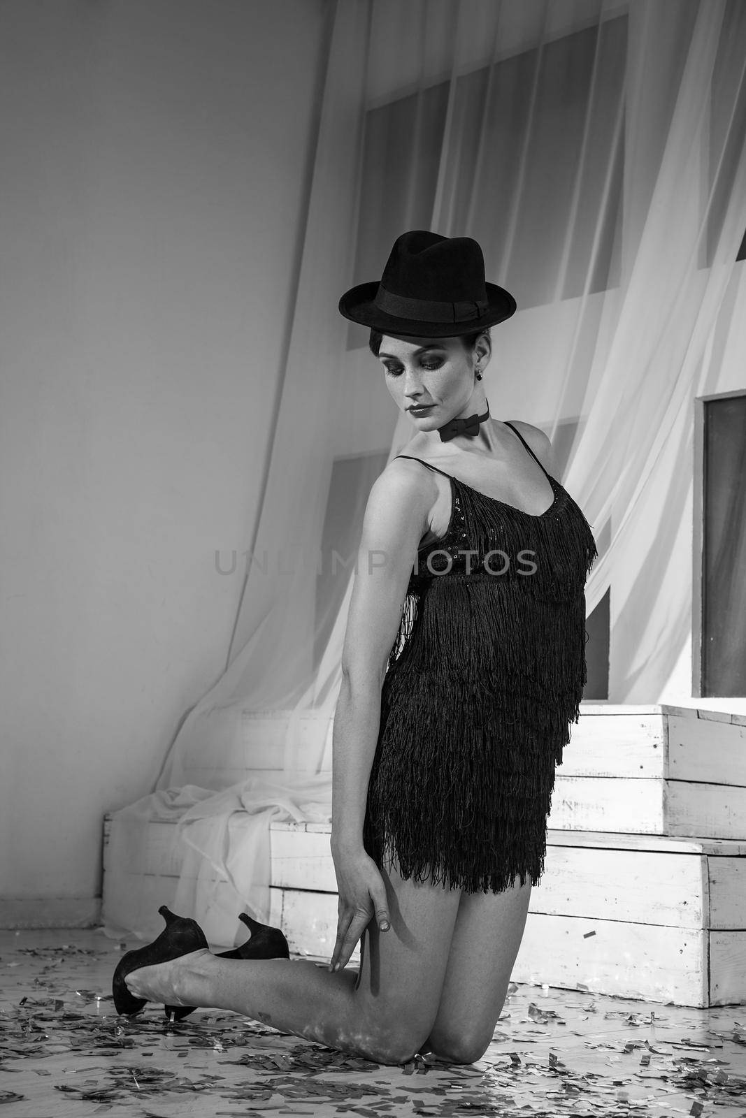 Beautiful young caucasian dancer in black dress and fashion makeup and hat posing standing on her knees and looking into the distance. Studio shot. Black and white photo.