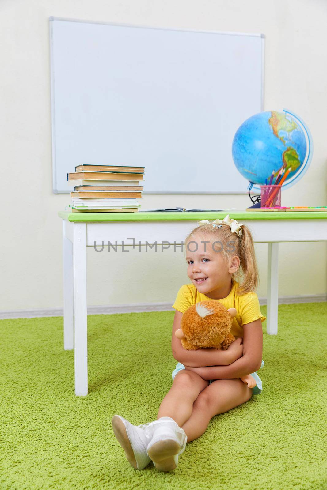 Portrait of cute little latin schoolgirl in classroom. Happy young latin girl wearing casual clothing sitting on a floor holding oy by Mariakray