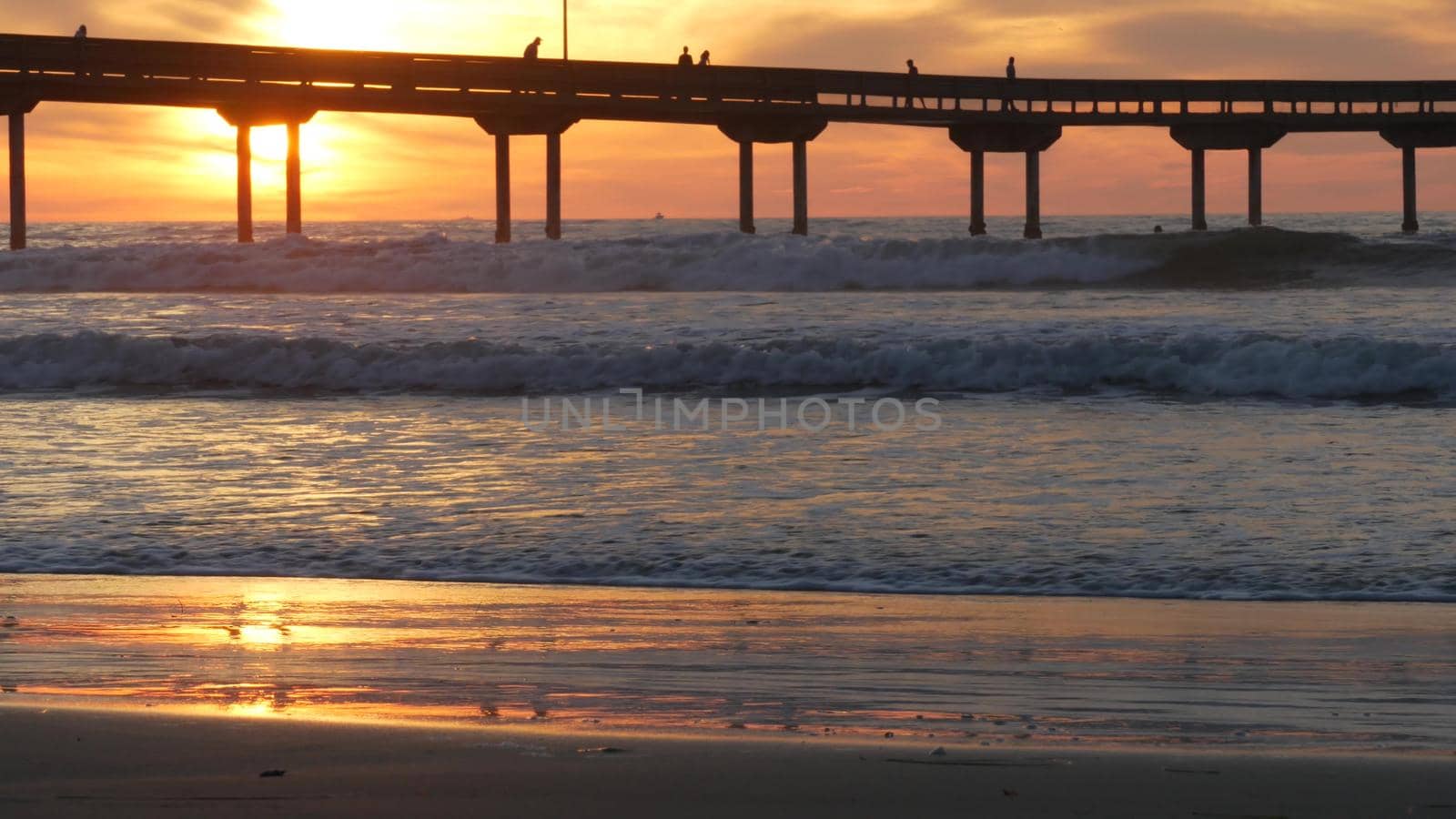 Silhouette of people walking, pier on piles in sea water. Ocean waves, dramatic sky at sunset. California coast aesthetic, beach or shore vibe at sundown. Summer seascape in San Diego near Los Angeles