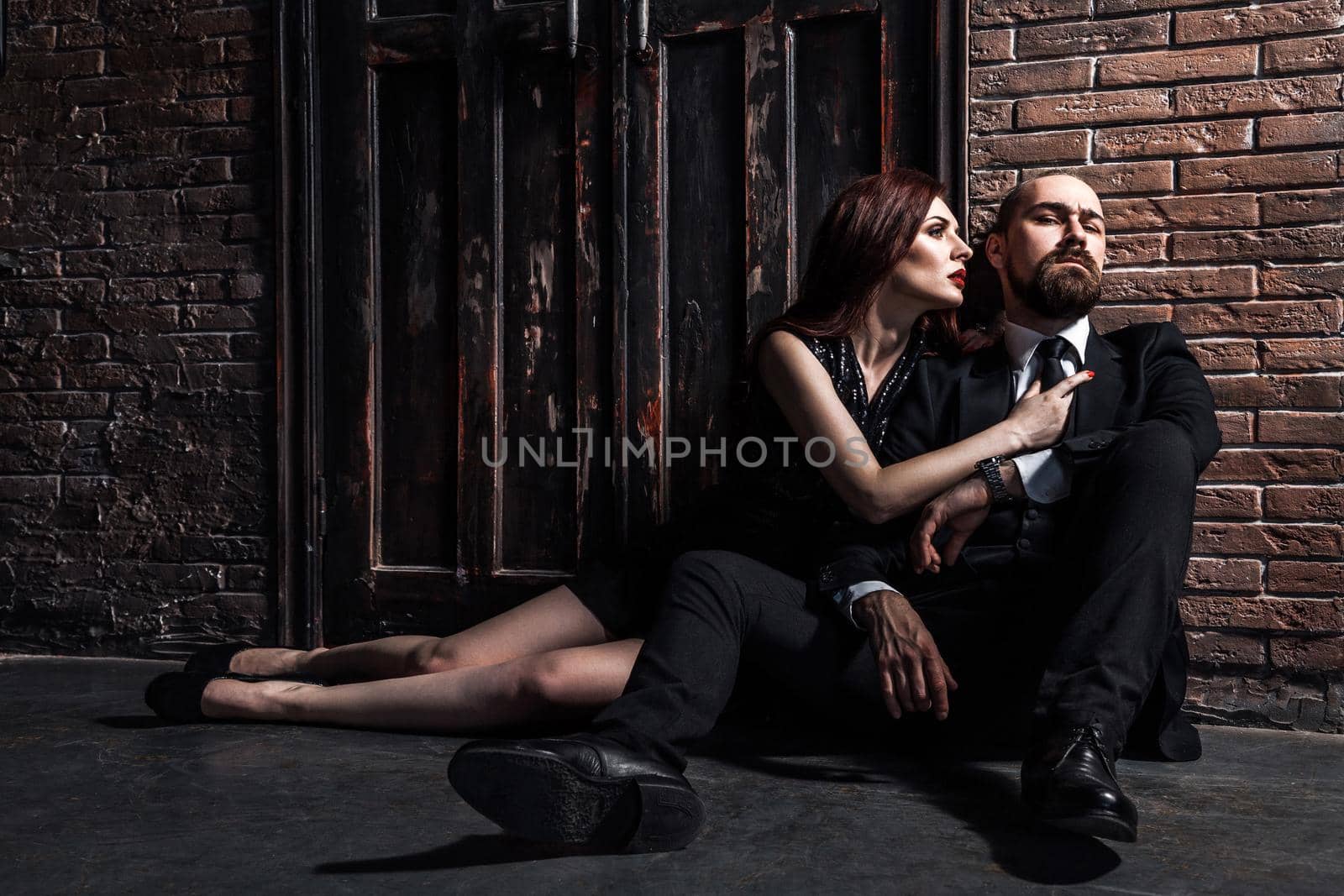 Man in suit and woman in evening dress sitting on his lap. Ginger woman looks at him and wants to kiss. Studio shot, brick wall