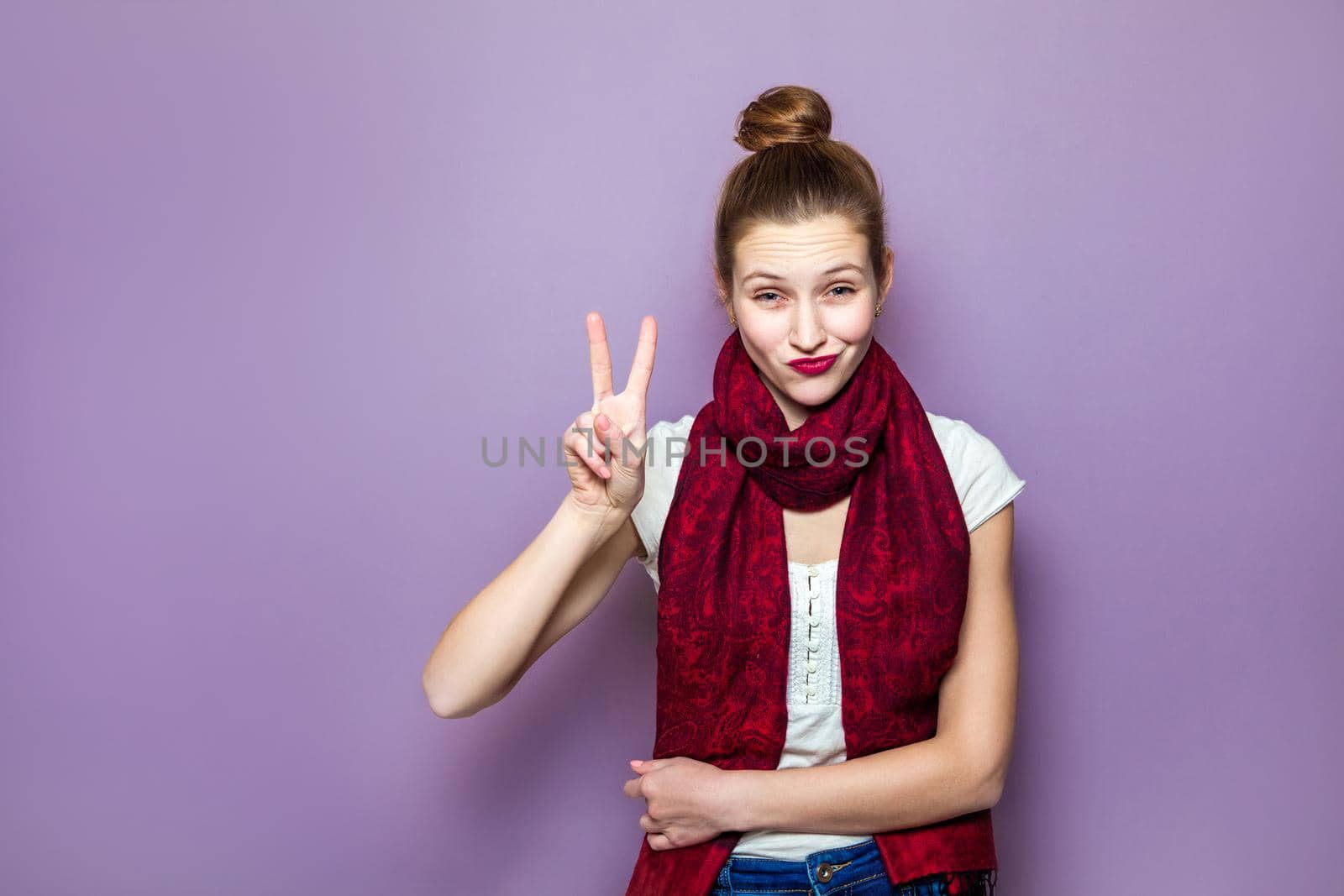 Portrait of beautiful young happy smiling woman, showing two fingers or victory gesture, dressed white t-shirt and red scarf on purple background.