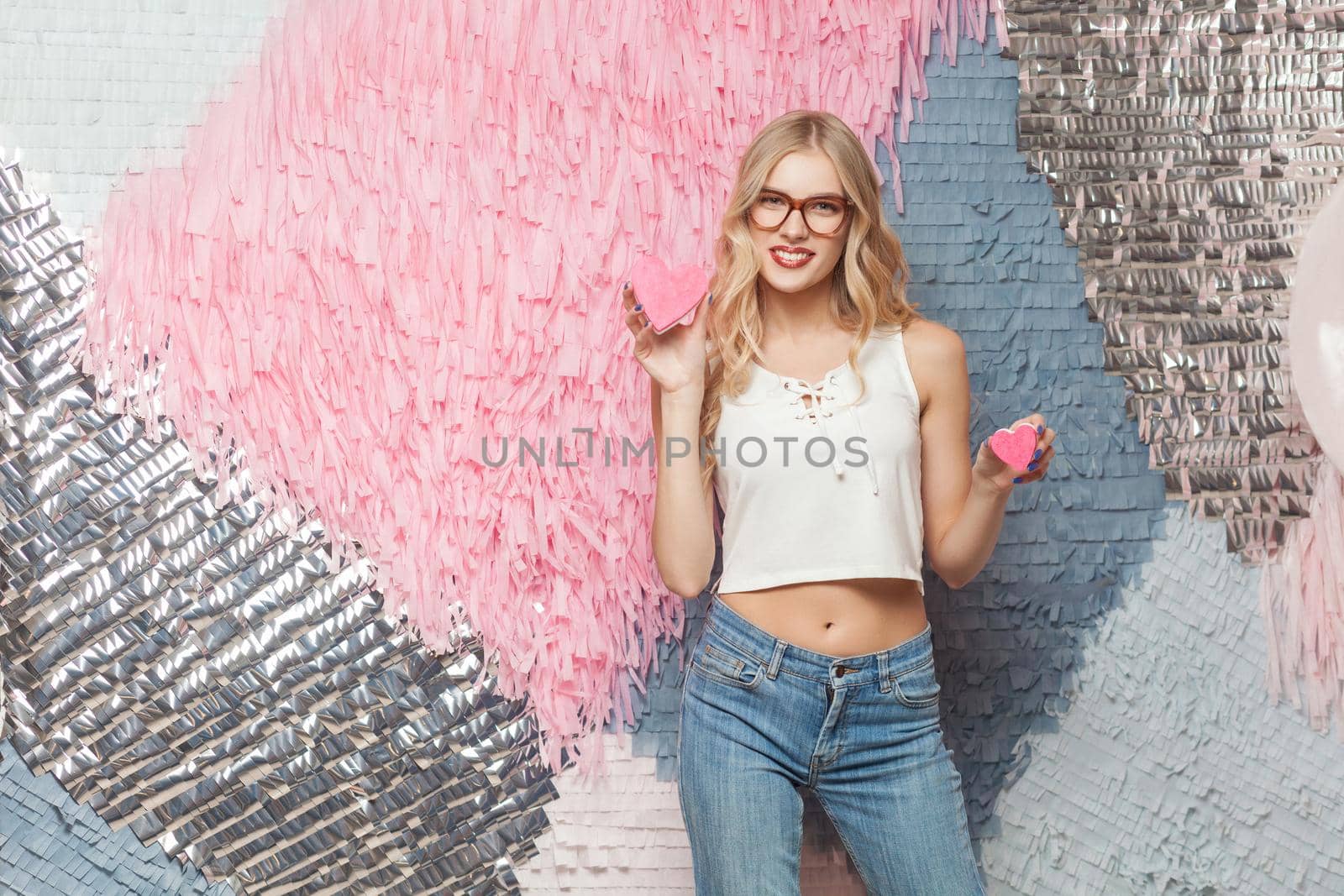 Beautiful young woman with blondie long hair in stylish glasses, looking at camera and holds two cookies in form of hearts. Isolated studio shot on colorful abstract background
