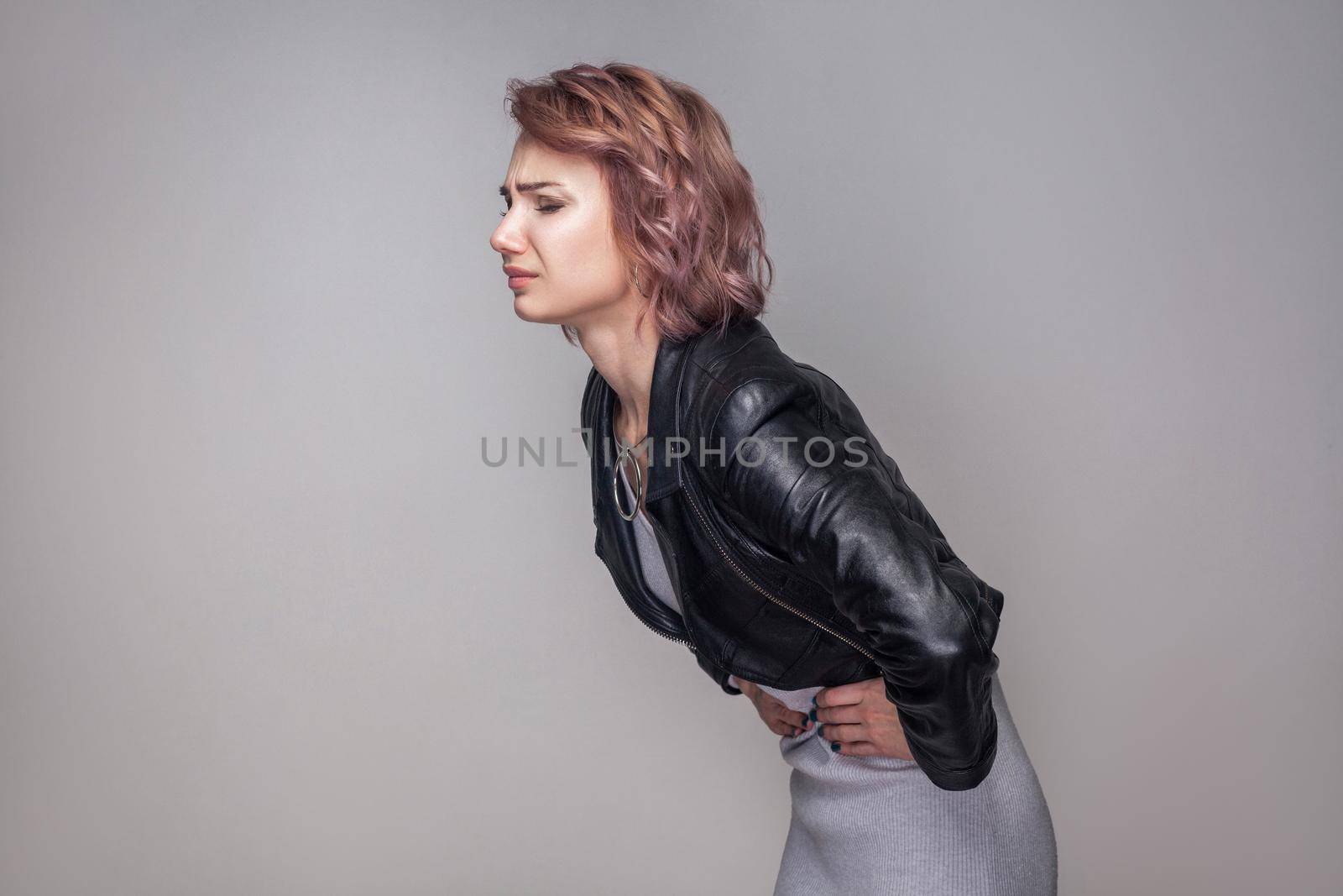Stomach pain. Profile side view portrait of woman with short hairstyle in casual style black leather jacket standing and holding her sick belly. indoor studio shot, isolated on grey background.