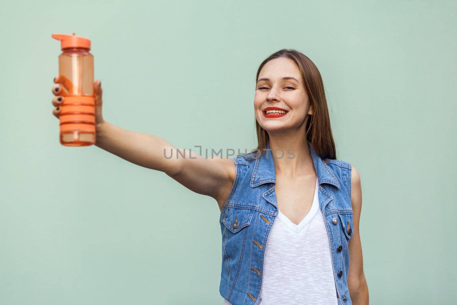 Young pretty woman with freckles in casual style show bottle arm forward and looking at camera with tooth smile face. Studio shot, isolated on a green background.