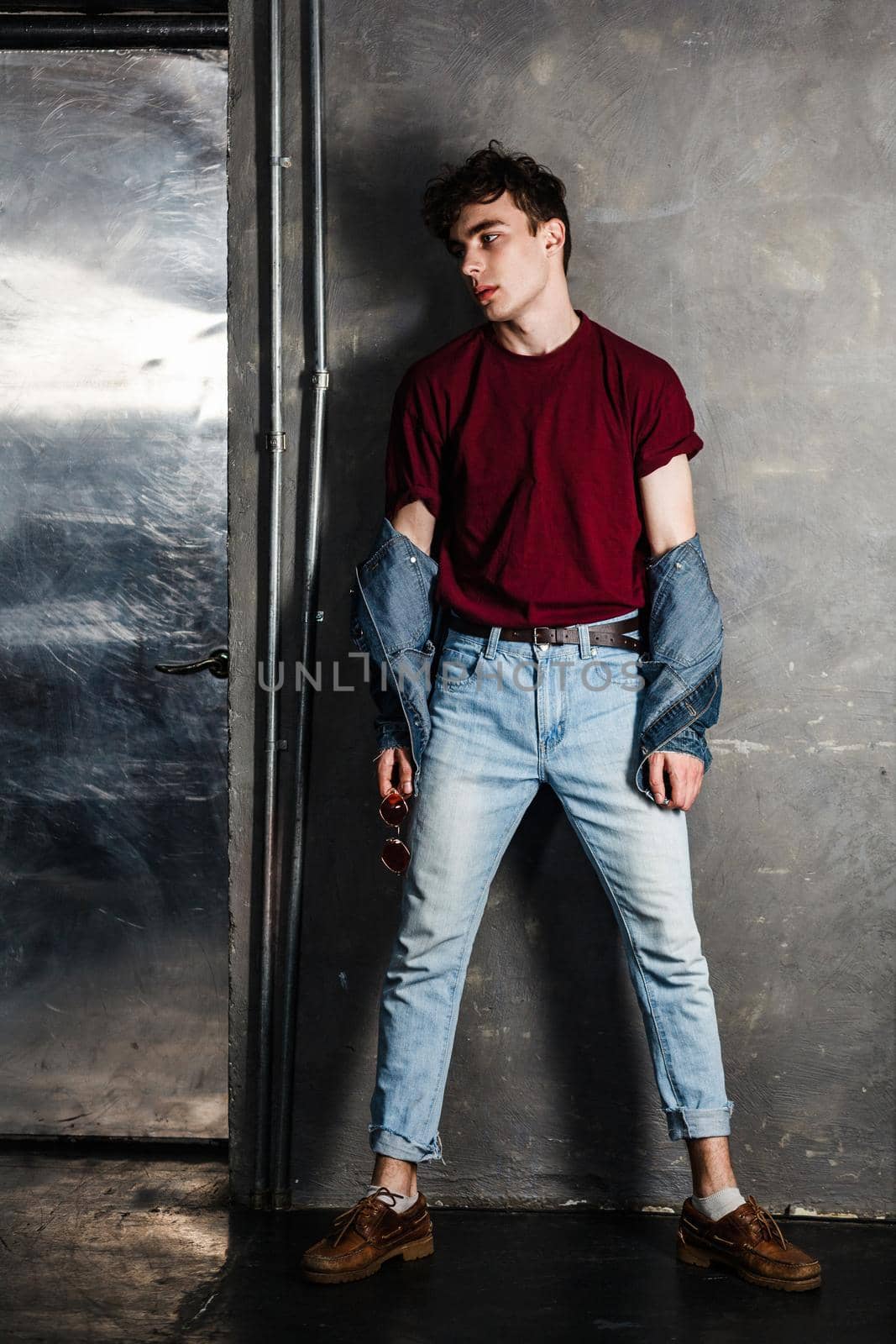 Full length portrait of stylish young fashion model man in bright red sunglasses and denim casual style posing near metallic door. standing and lean on grey wall and looking away. indoor, studio shot.