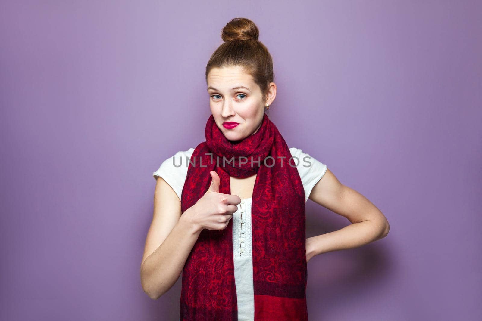 Thumbs up, young emotional girl with collected hair, freckles and red scarf looking excited with thumbs up on purple background, showing her thumb finger with happy funny face, looking at camera. .