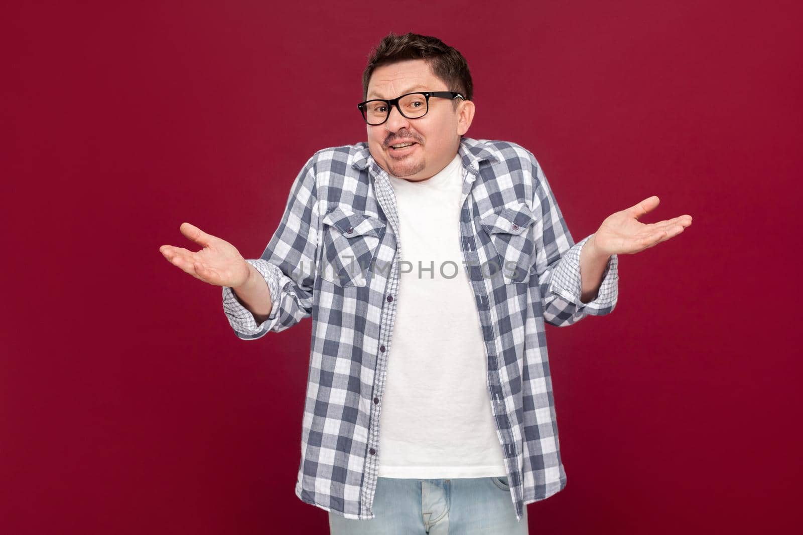I don't know. Portrait of confused handsome middle aged man in casual checkered shirt and eyeglasses standing with raised arms and dont know what to do. studio shot, isolated on dark red background.