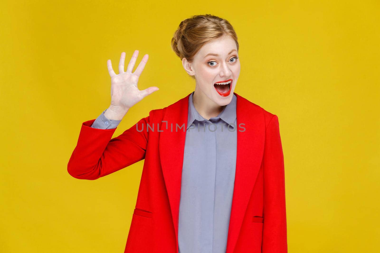 Hello! Ginger red head business woman in red suit showing hi sign. Studio shot, isolated on yellow background