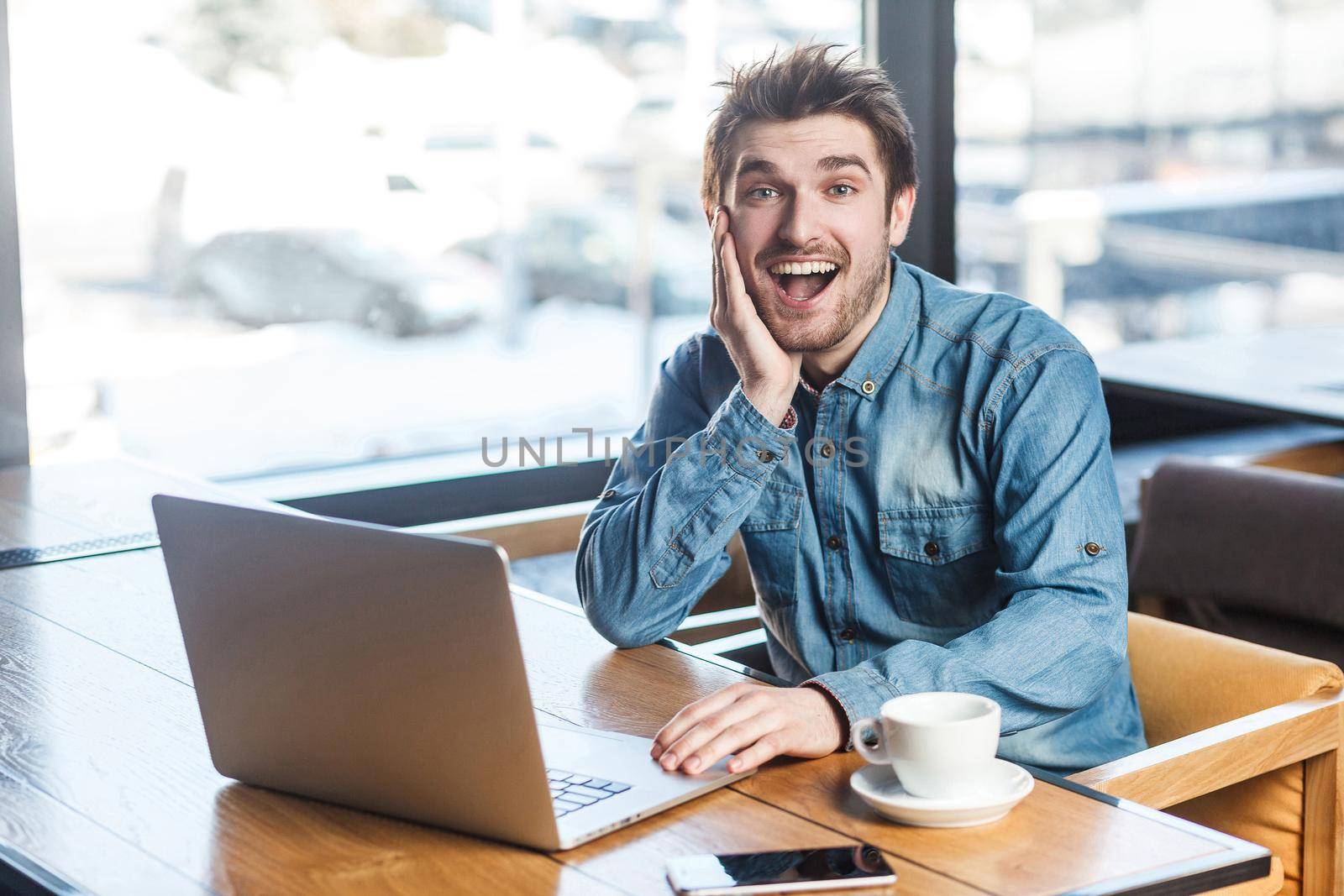 Oh my God! Portrait of happy toothy smiling bearded young freelancer in blue jeans shirt are sitting alone in cafe and having new proposal for a work on laptop are in positive mood, looking at camera.