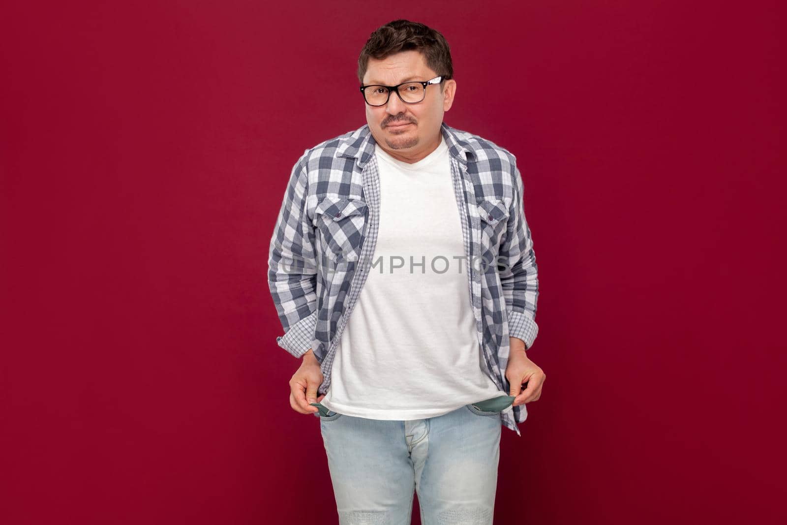 I don't have money more. Portrait of poor sad middle aged man in casual checkered shirt and eyeglasses standing looking and showing his empty pocket. indoor studio shot isolated on dark red background