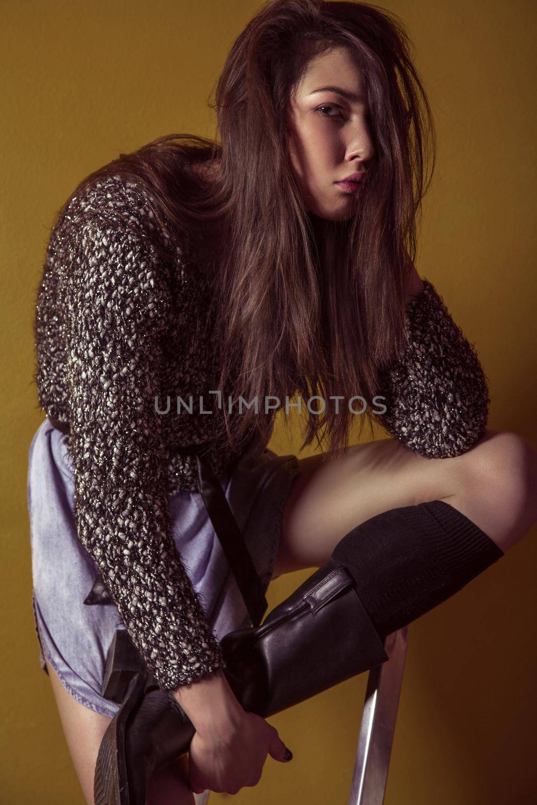 Beautiful young asian model in blue denim skirt and black blouse and fashion makeup posing sitting on stepladder and looking at camera. Studio shot. High-fashion photo.