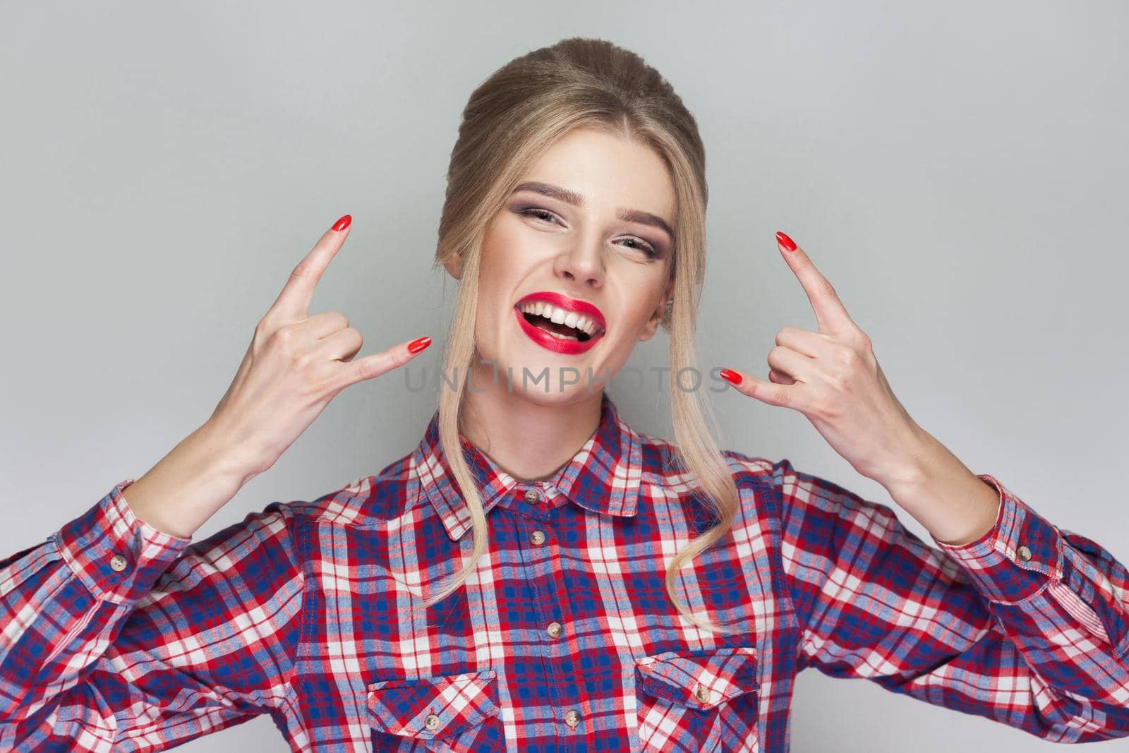 funny kissing beautiful blonde girl with pink checkered shirt, collected updo hairstyle and makeup standing and showing Ok sign and looking at camera with wink. indoor , isolated on gray background.