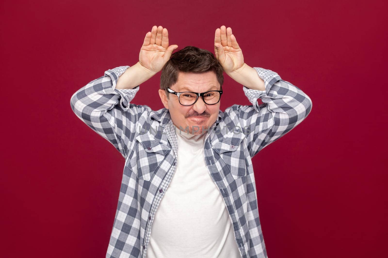 Portrait of funny handsome middle aged business man in casual checkered shirt and eyeglasses standing with bunny ears gesture on head, looking at camera. studio shot, isolated on dark red background.