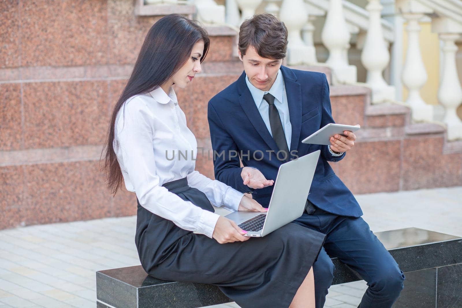 Boss in blue suit and his assistant in white blouse and black skirt discussing their work outdoors.