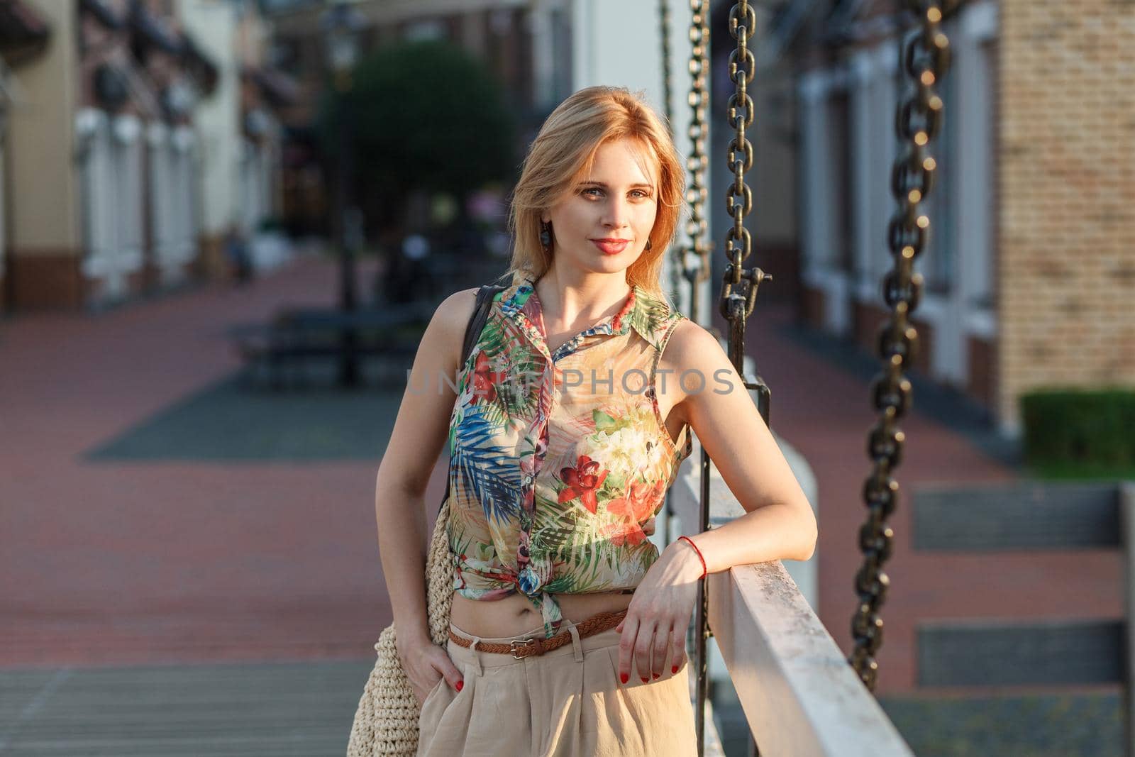 Beautiful young woman in tropical shirt, beige galligaskins with wicked bag standing with hand in pocket and posing on city background and looking at camera with smile in summertime. outdoor shot.