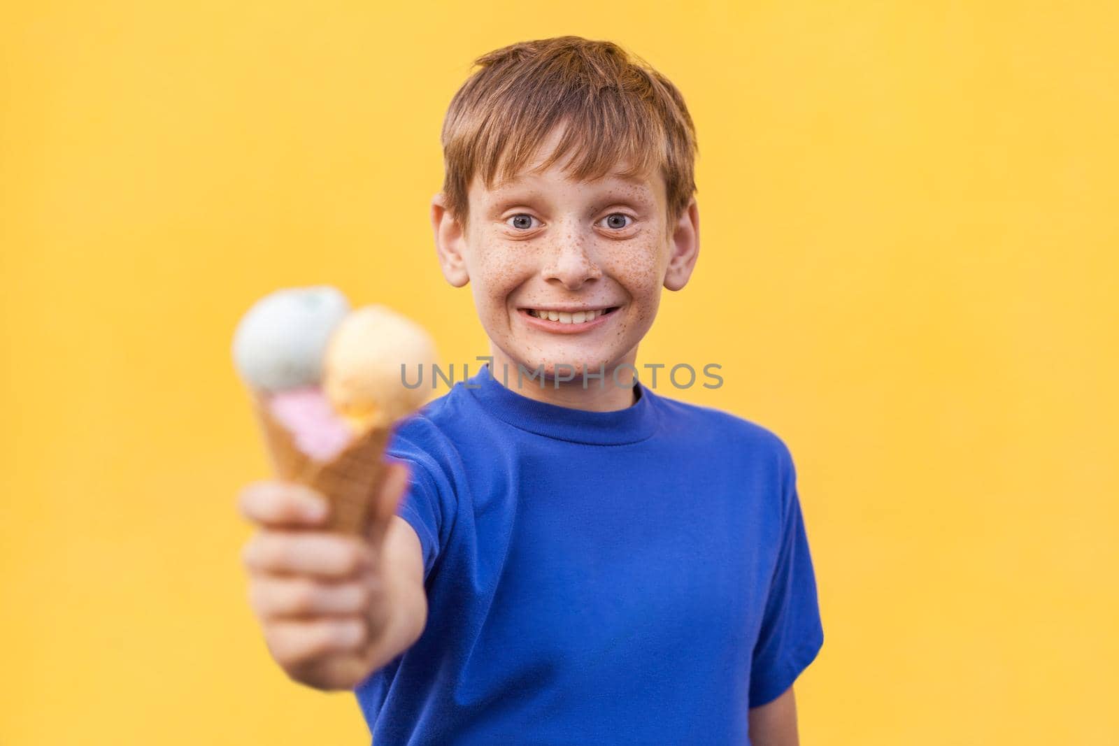 Blonde beautiful boy with freckles and blue T-shirt showing ice cream and looking at camera with big eyes and toothy smile. Studio shot, isolated on a yellow background.