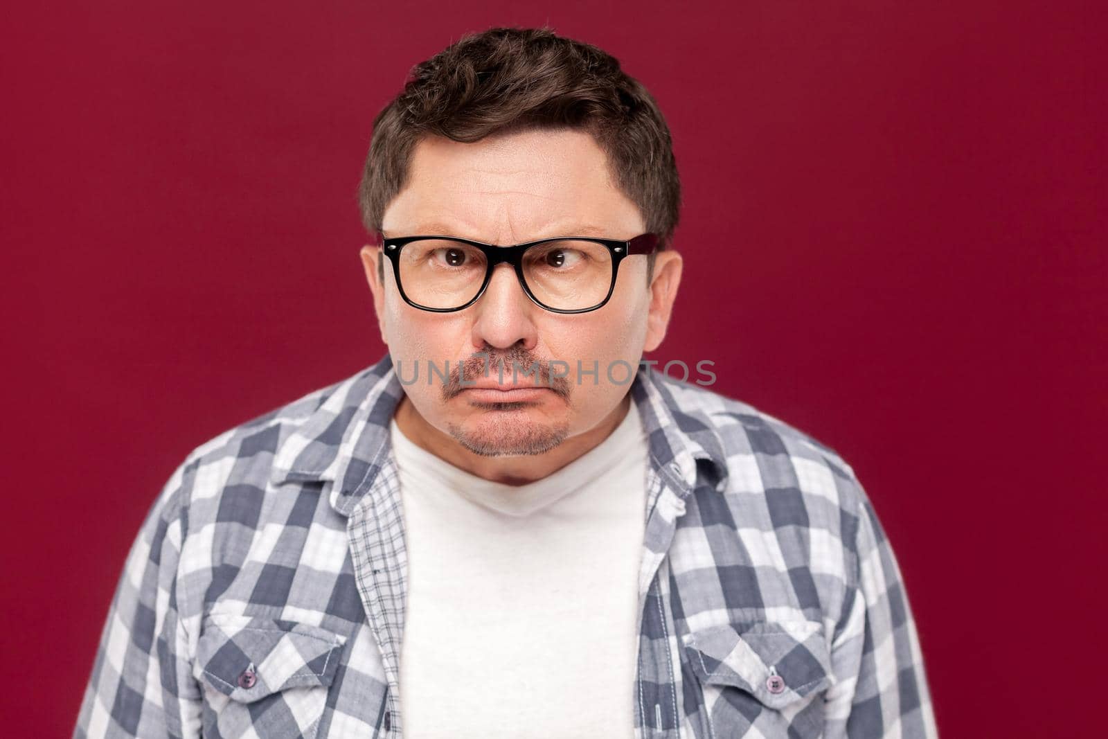 Closeup portrait of crazy funny middle aged business man in casual checkered shirt, eyeglasses and mustache standing with crossed eyes and looking. indoor studio shot, isolated on dark red background.