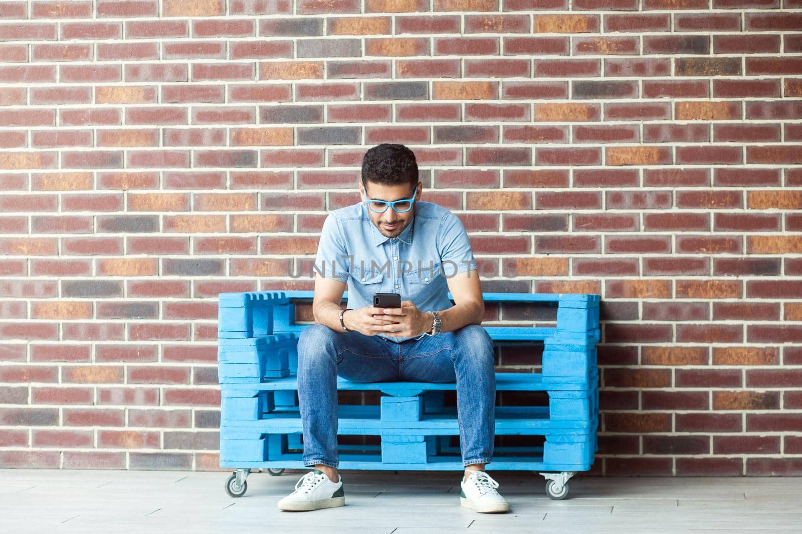 Full length portrait of successful handsome young bearded man in casual style, eyeglasses sitting on blue wooden pallet, holding, looking at mobile smartphone. indoor studio shot on brown brick wall.