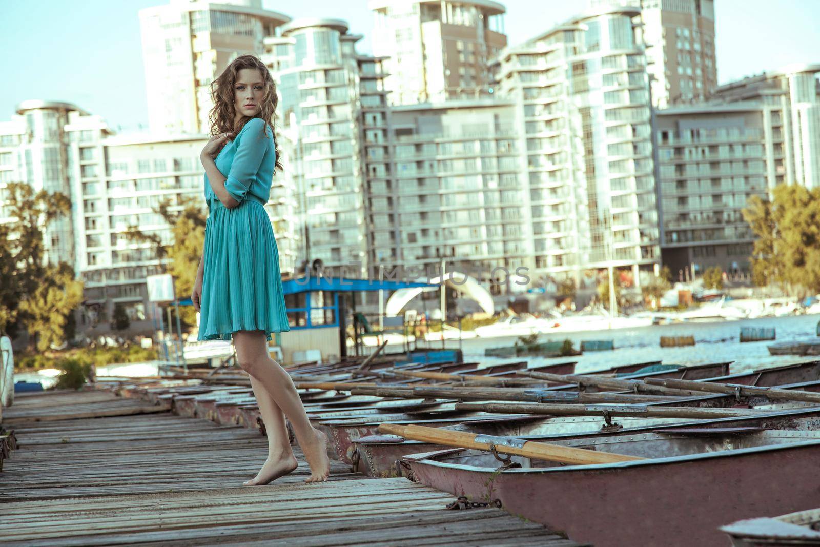 Beautiful young fashion model in green turquois dress and fashion makeup and hairstyle is posing on pier, posing and looking at camera on city background.