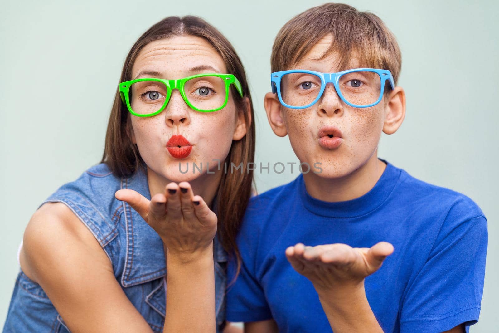 Young sister and brother with freckles on their faces, posing over light blue background together, looking at camera and sending kisses. Studio shot