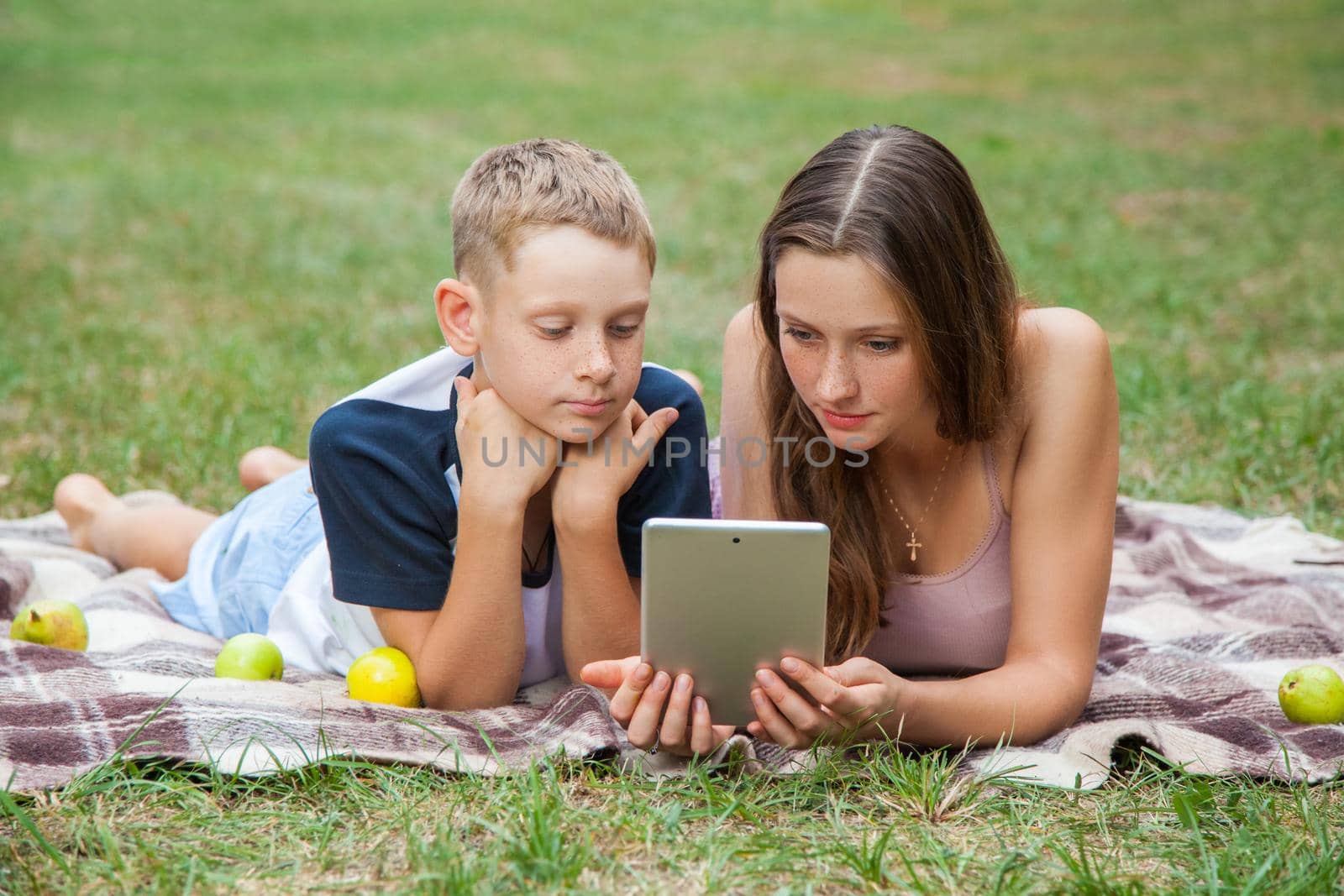 Young sister and brother with freckles on their faces lying down on plaid and using tablet in park. looking at tablet display, top view.