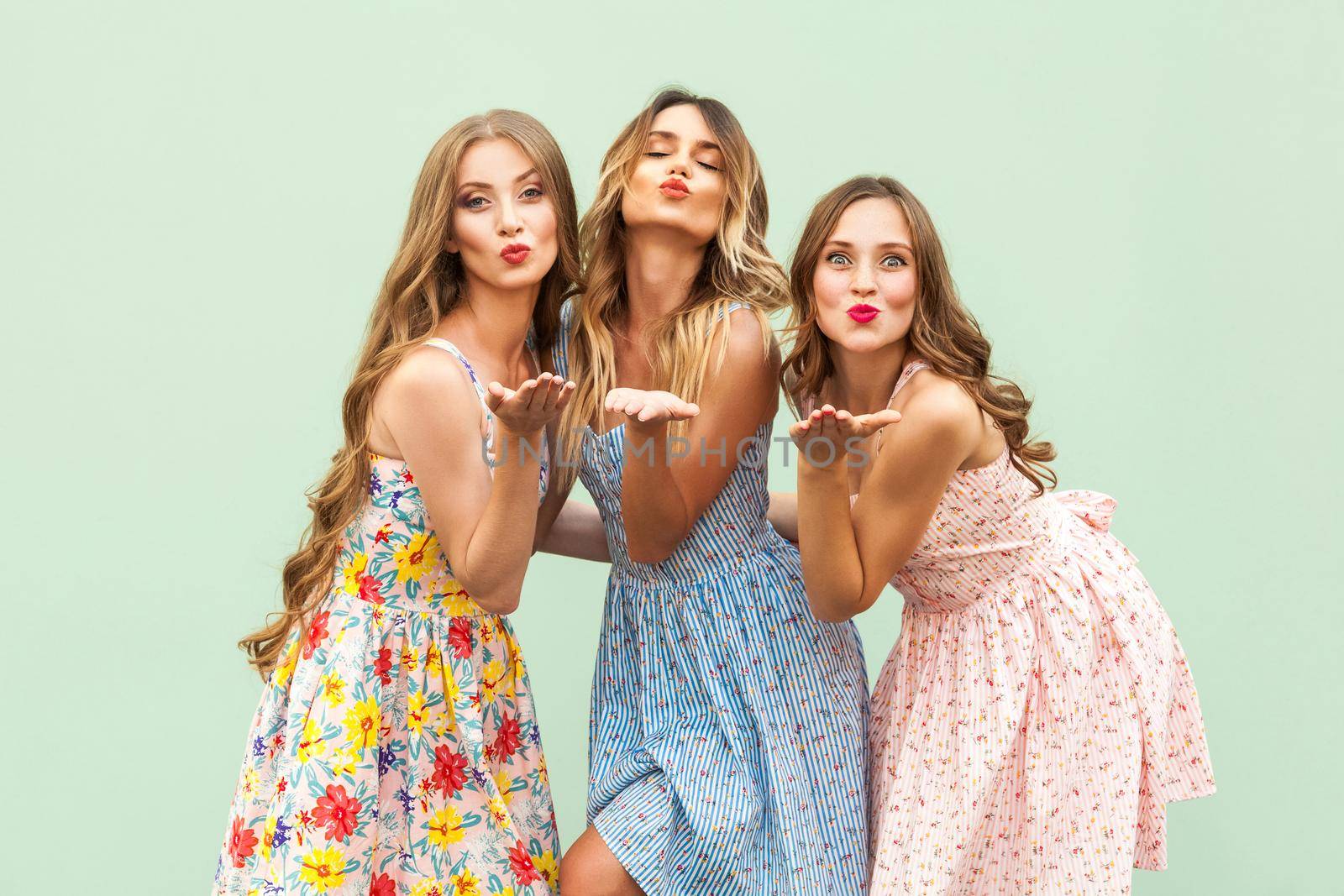 Sending air kiss. Three best friends posing in studio, wearing summer style dress against green background. by Khosro1