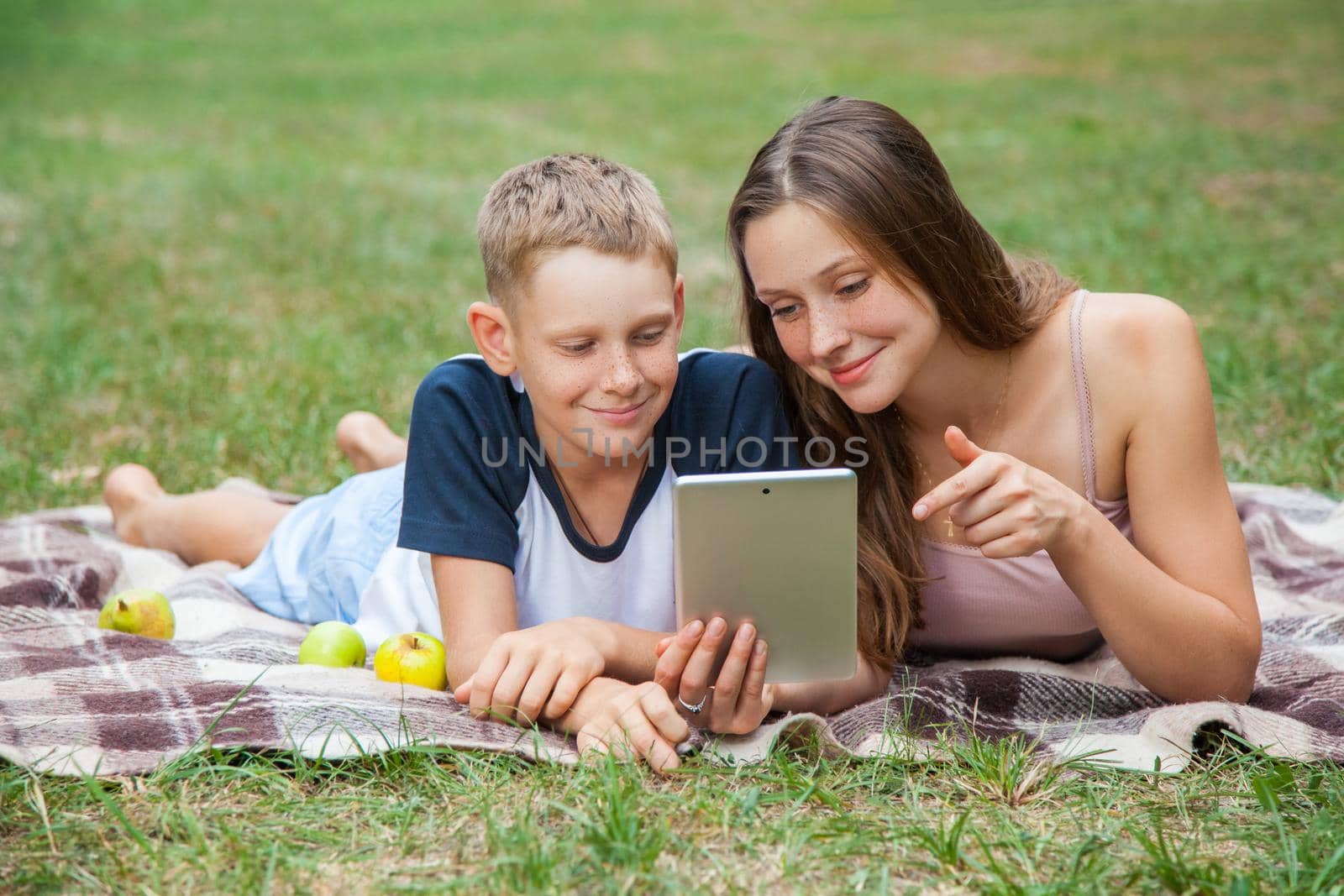 Young sister and brother with freckles on their faces lying down on plaid and using tablet in park. she is showing display with her finger and smiling.