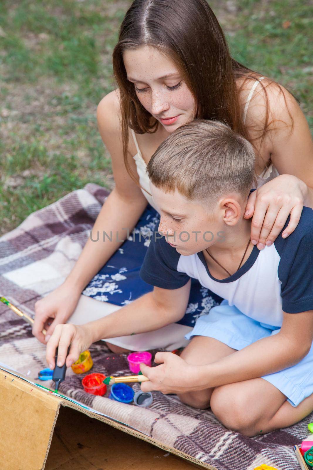 Older sister trying to teach her brother to paint and posing in the park on summer time, drawing and painting..