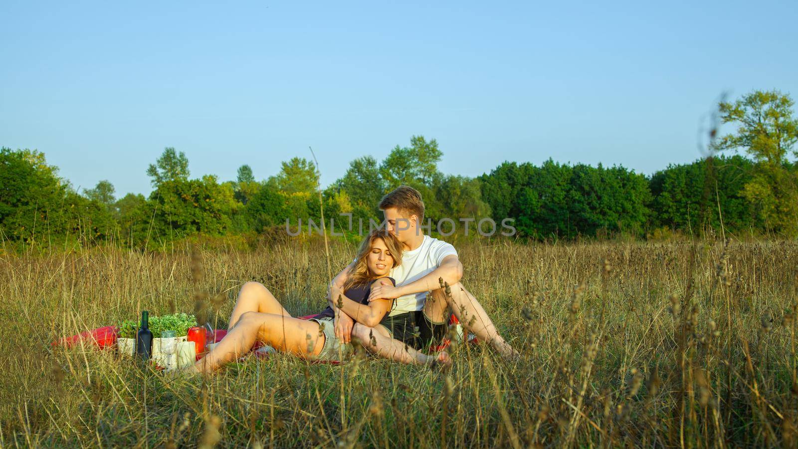 Beautiful young happy loving couple on picnic lying down on sunny summer day enjoying and resting. he is kissing with closed eyes.