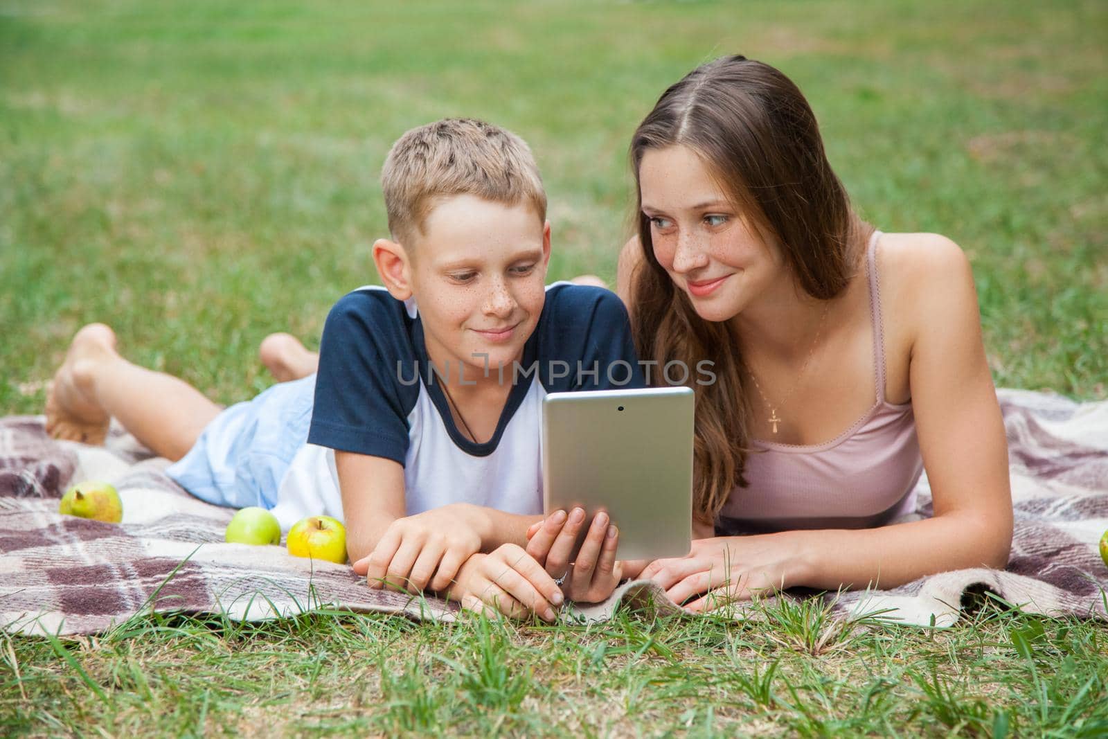 Young sister and brother with freckles on their faces lying down on plaid and using tablet in park. looking at tablet and smiling.