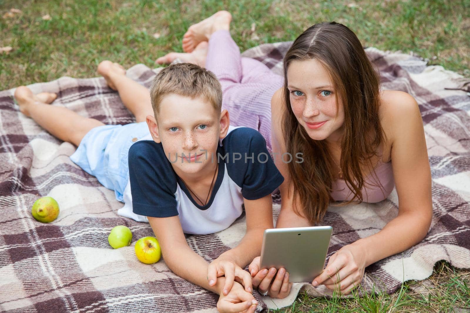 Young sister and brother with freckles on their faces lying down on plaid and using tablet in park. top view, looking at camera with smile.