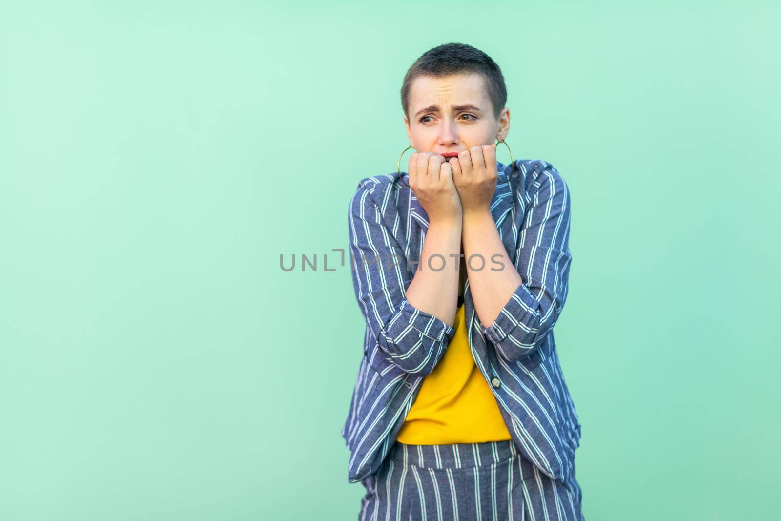 Portrait of nervous handsome beautiful short hair young stylish woman in casual striped suit standing, bitting her nails and panic. indoor studio shot isolated on light green background.