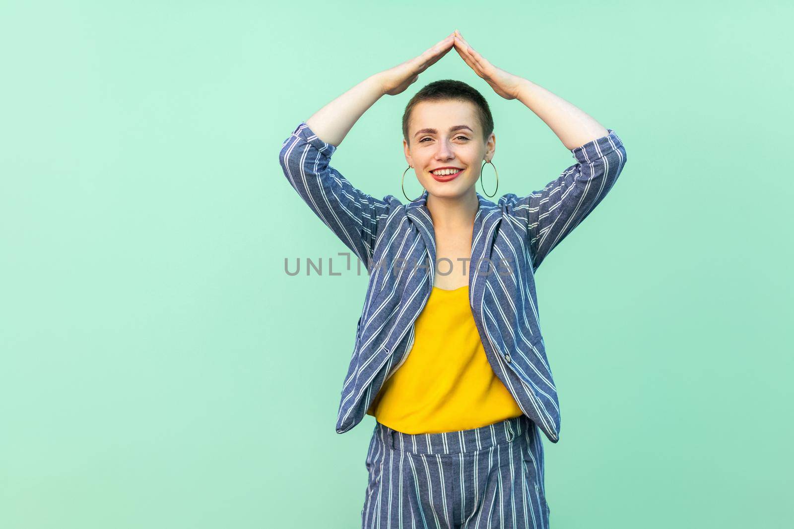 Portrait of happy handsome beautiful short hair young stylish woman in casual striped suit standing with roof gesture on head with hands, toothy smile. studio shot isolated on light green background.