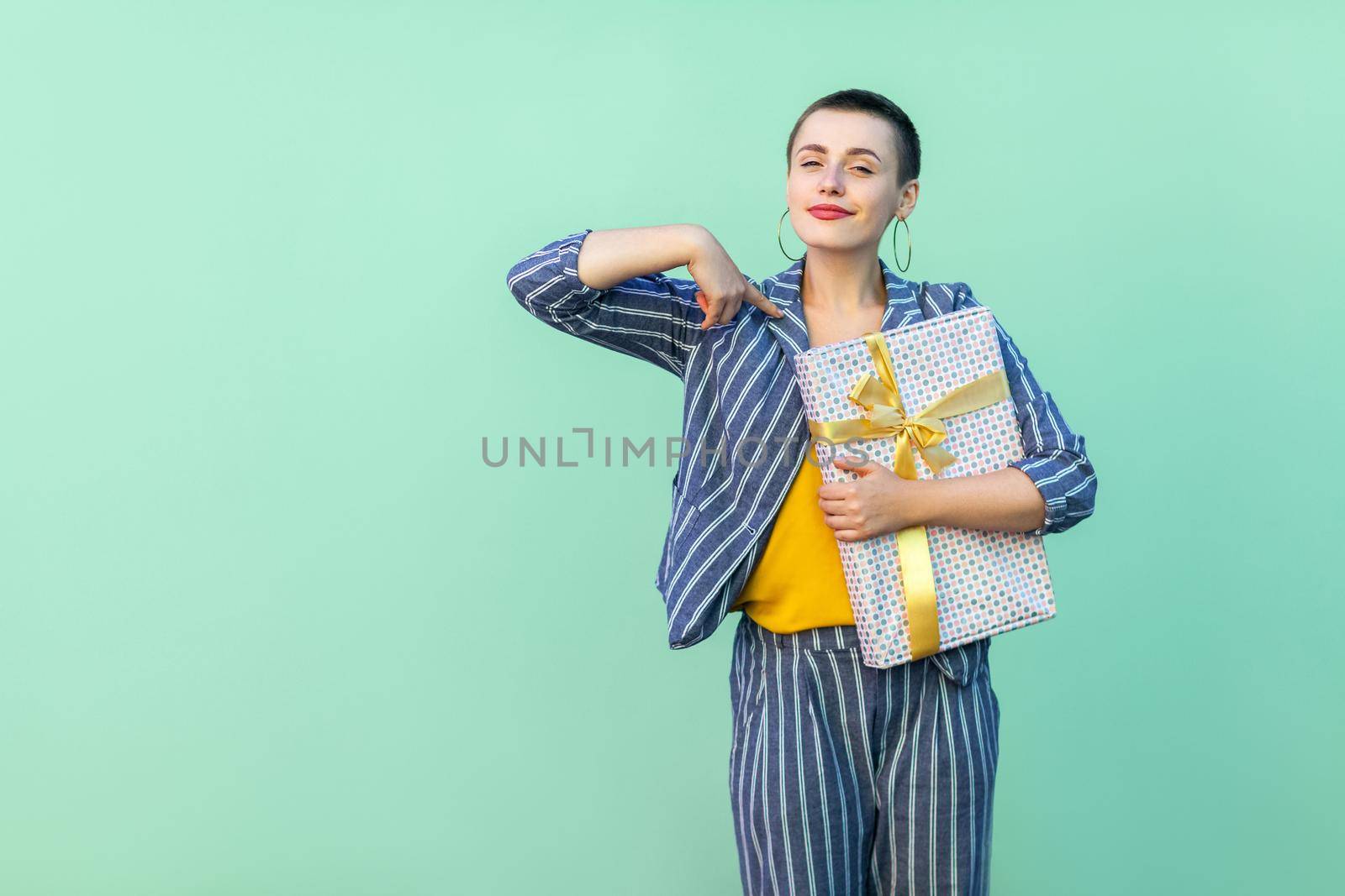 Portrait of confident beautiful with short hair young woman in striped suit standing, pointing finger to herself and holding present box with proud face. Indoor, isolated,studio shot, green background