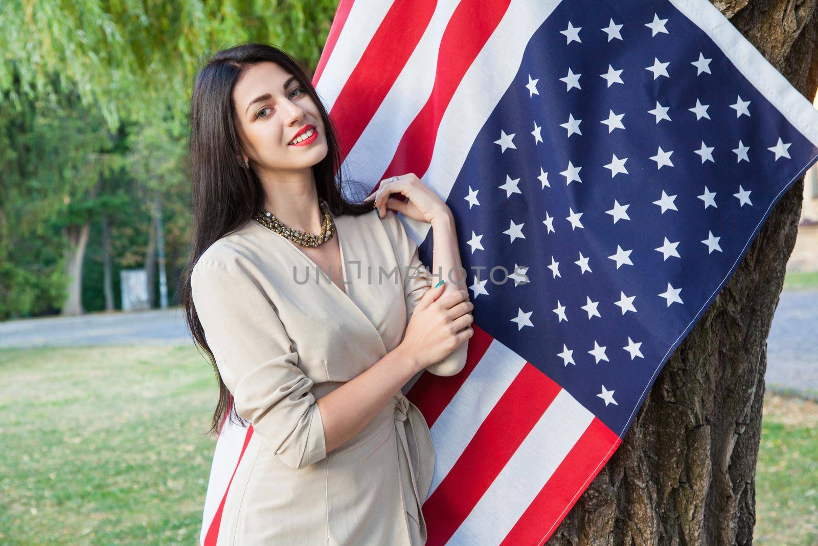 Beautiful young woman with classic dress near american flag in the park. fashion model holding us smiling and looking at camera. usa lifestyle. posing near tree and toothy smile.