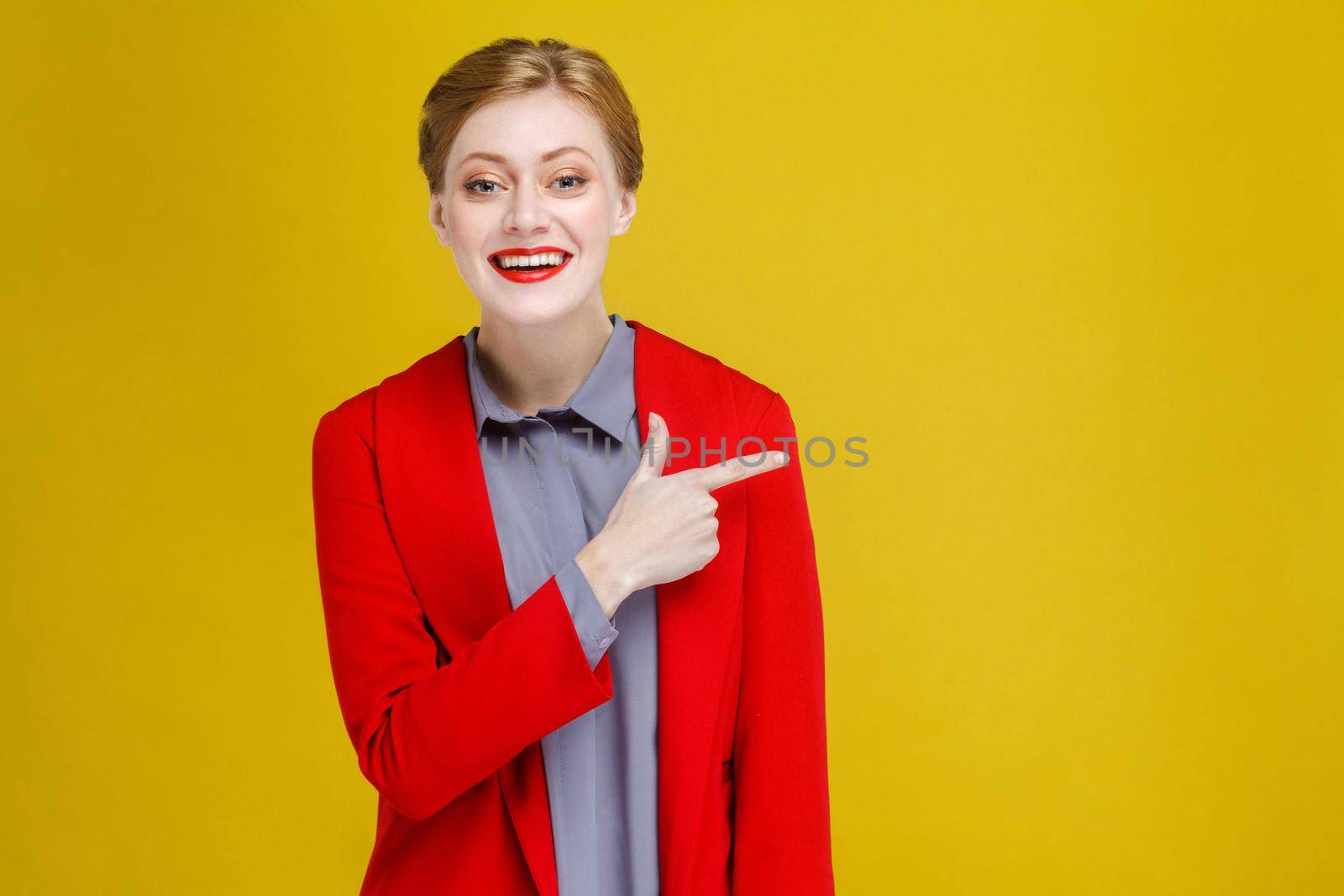 Redhead business woman in red suit showing at copy space. Studio shot, isolated on yellow background