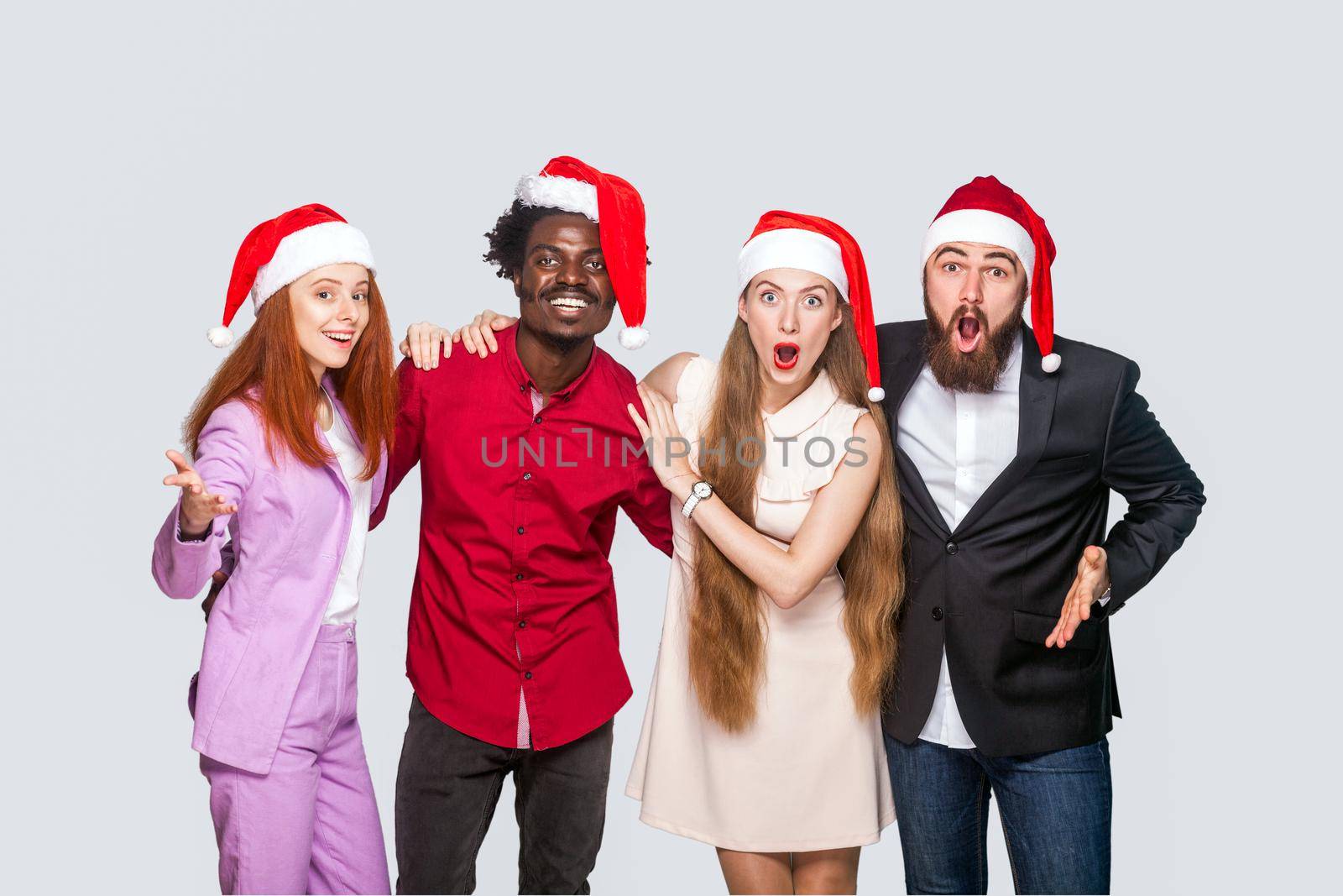 Group of beautiful happy handsome friends in red cap standing and celebrating new year and looking at camera with amazed surprised face. indoor studio shot, isolated on gray background.