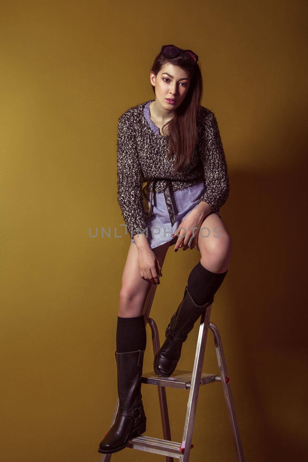 Beautiful young asian model in blue denim skirt and black blouse and fashion makeup posing sitting on stepladder and looking at camera. Studio shot. High-fashion photo.