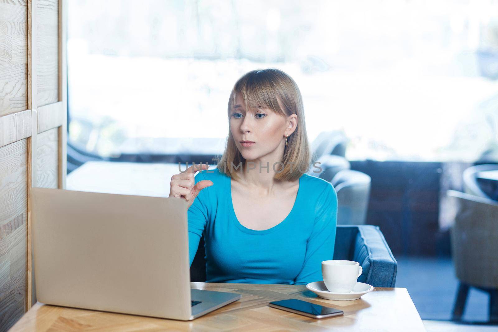 Little bit! Portrait of attractive young girl with blonde hair in blue blouse are sitting in cafe, working and making video call on laptop, pleased her companion to give more time with pretty face