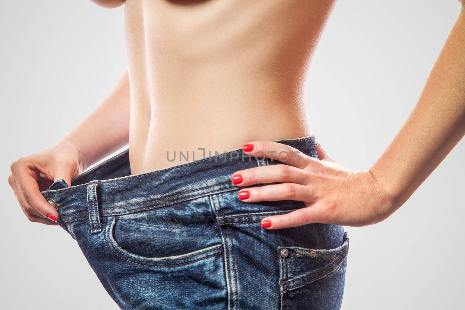 Closeup of slim waist of young woman standing in big jeans showing successful weight loss, indoor studio shot, isolated on light gray background, diet concept.