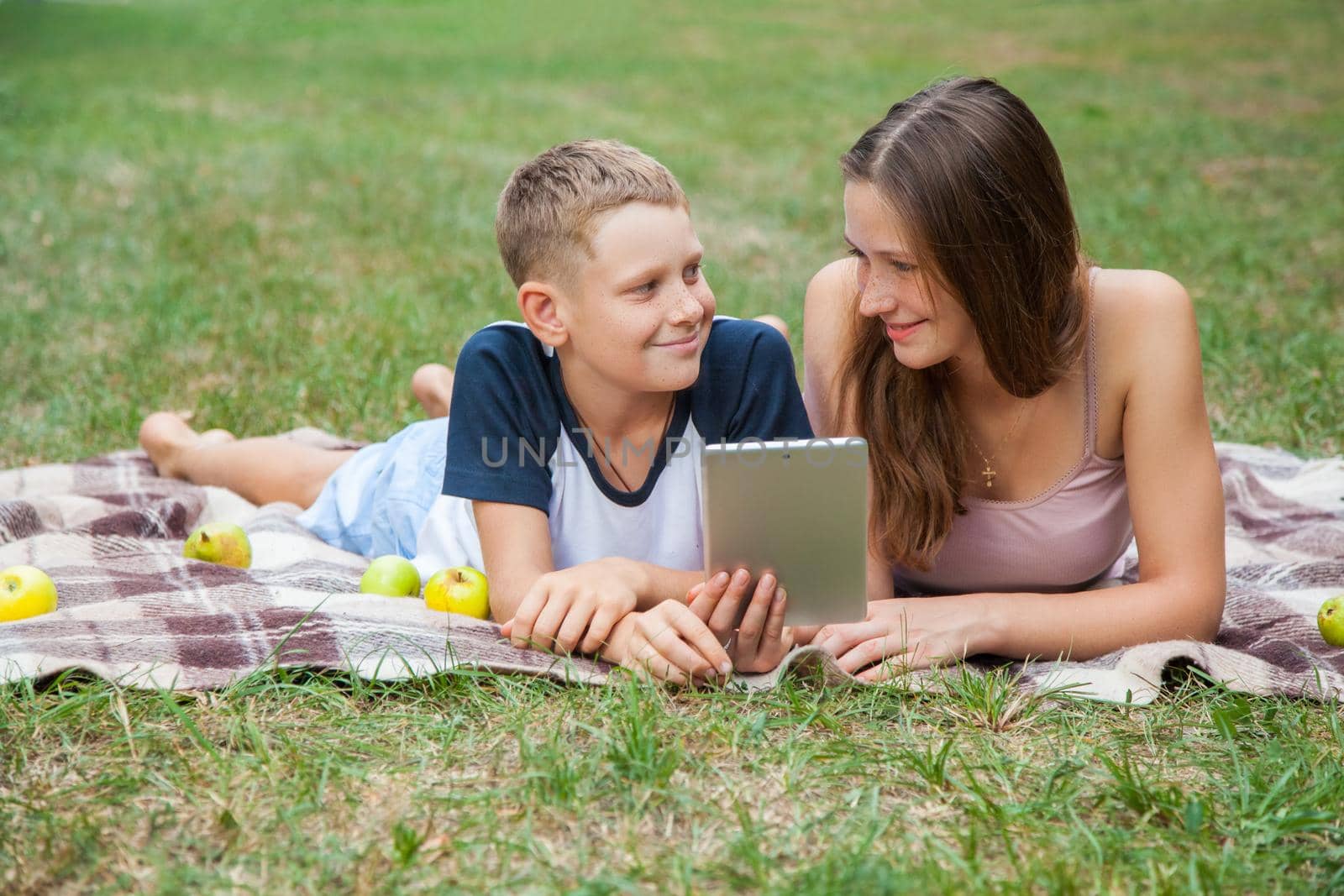 Young sister and brother with freckles on their faces lying down on plaid and using tablet in park. looking to each other and smiling.
