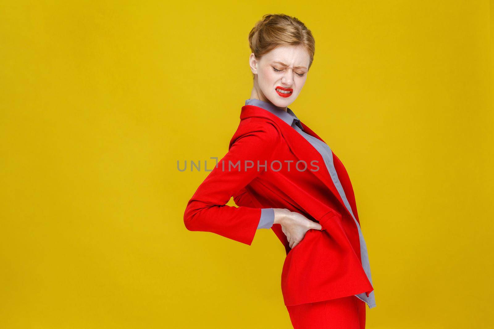 Redhead business woman in red suit have kidneys pain or back. Studio shot, isolated on yellow background