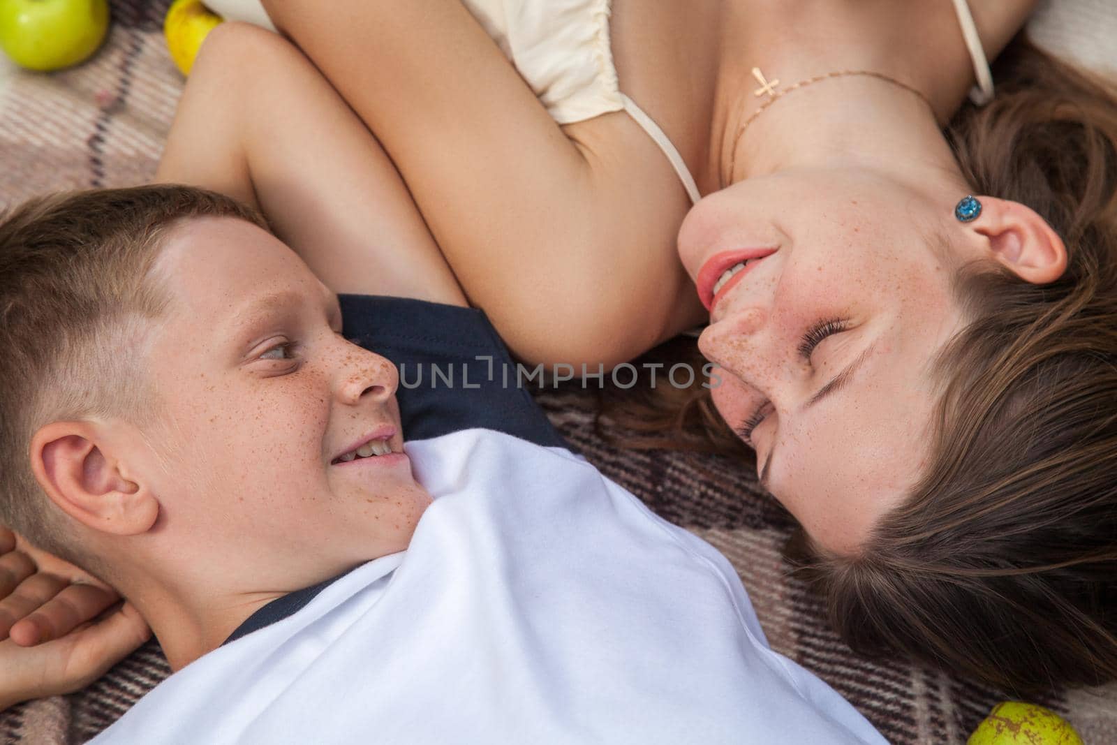 Happy young beautiful sister and brother with freckles on their faces posing lying down on plaid and smiling and looking at each other with toothy smile.