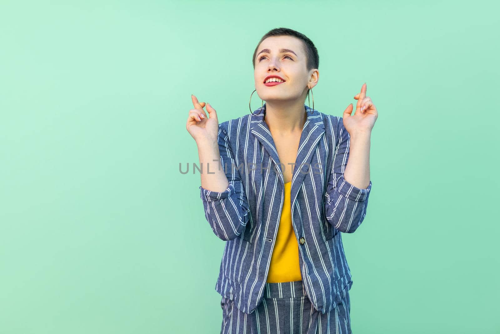Portrait of hopeful handsome beautiful short hair young stylish woman in casual striped suit standing, crossed fingers and wishful to win . indoor studio shot isolated on light green background.