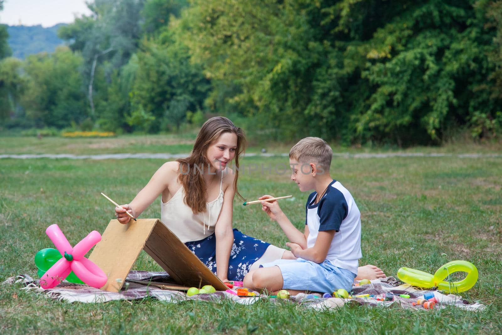 Older sister trying to teach her brother to paint and posing in the park on summer time, looking and smiling.