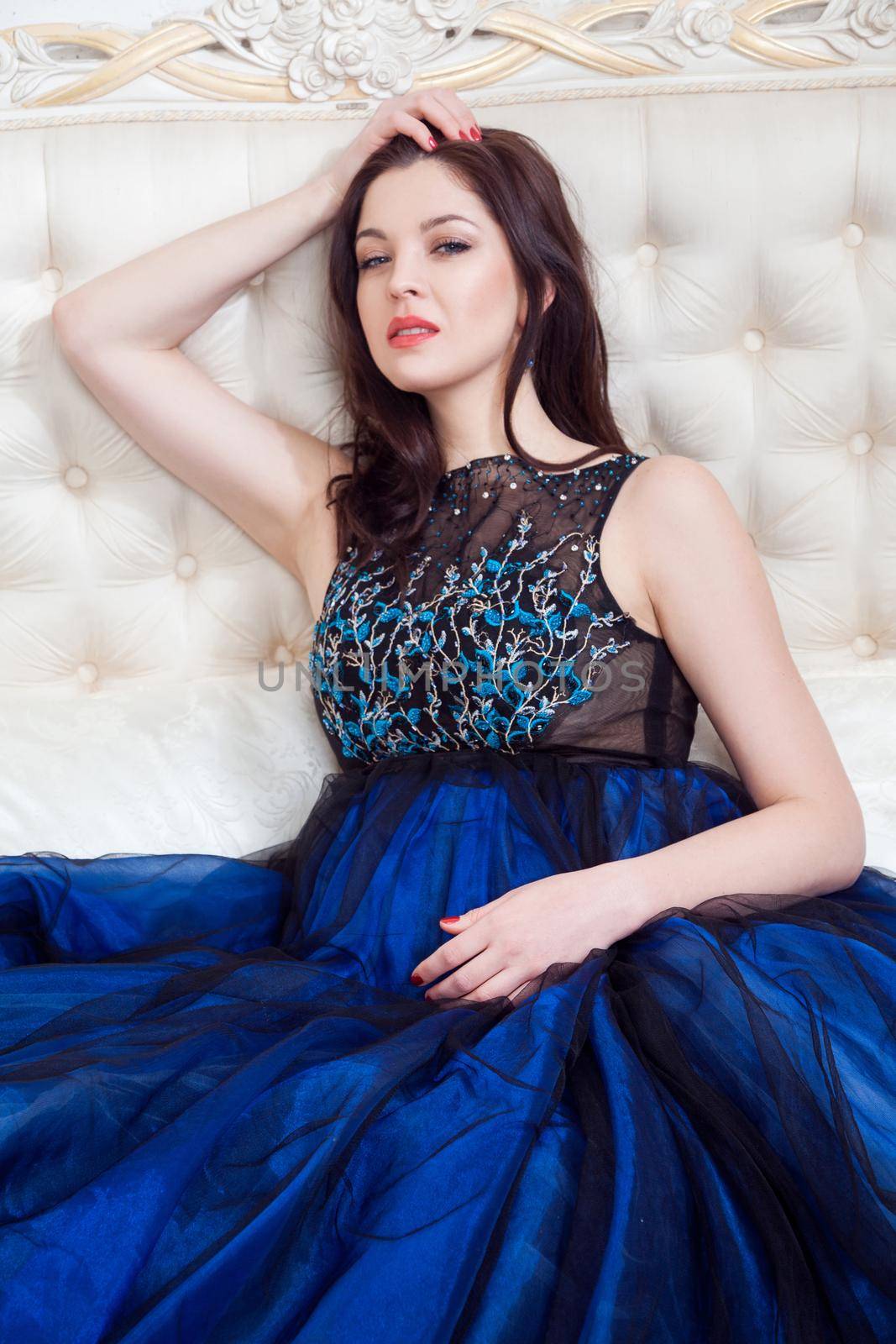 Attractive brunette woman with beautiful makeup in blue evening dress posing while sitting on bed, holding hand on head and looking at camera with pasion. indoor studio shot.